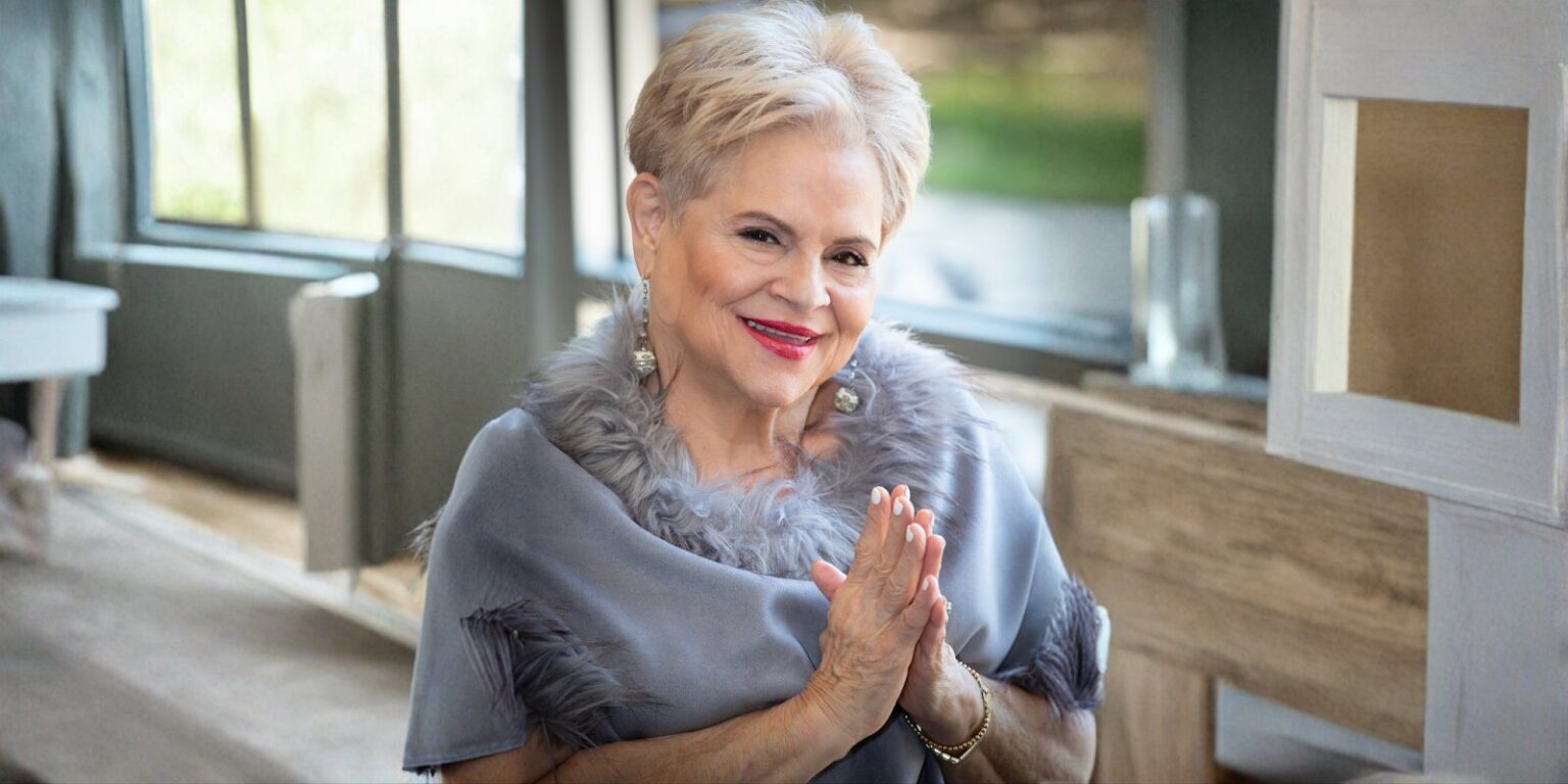 Elaine Adita Agnes smiling warmly, dressed in a chic, fur-trimmed gray shawl, radiating confidence and authenticity.