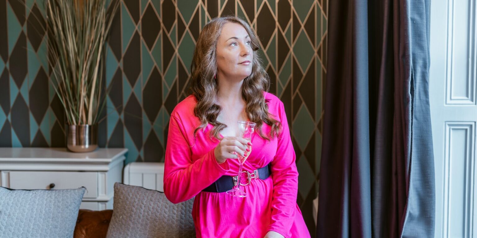 A woman with long wavy hair in a pink dress holding a glass, gazing thoughtfully by a window with a potted plant.
