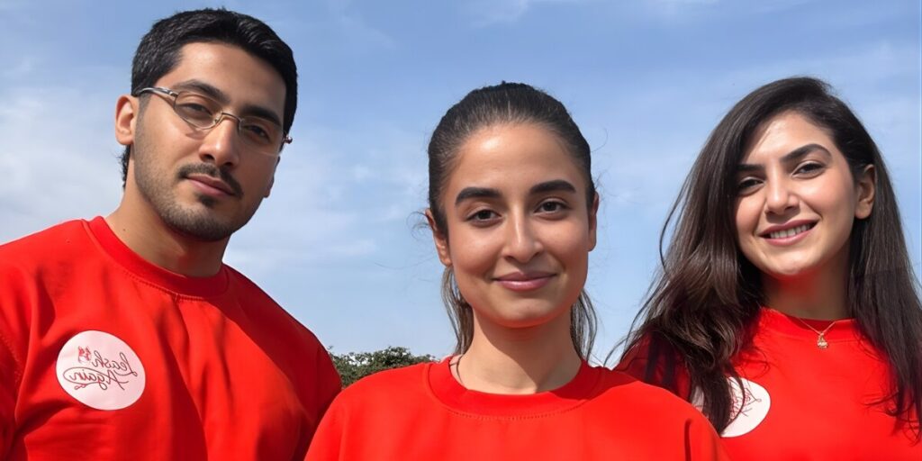 three smiling individuals wearing red LeashAgain shirts, standing together outdoors with a bright sky in the background.