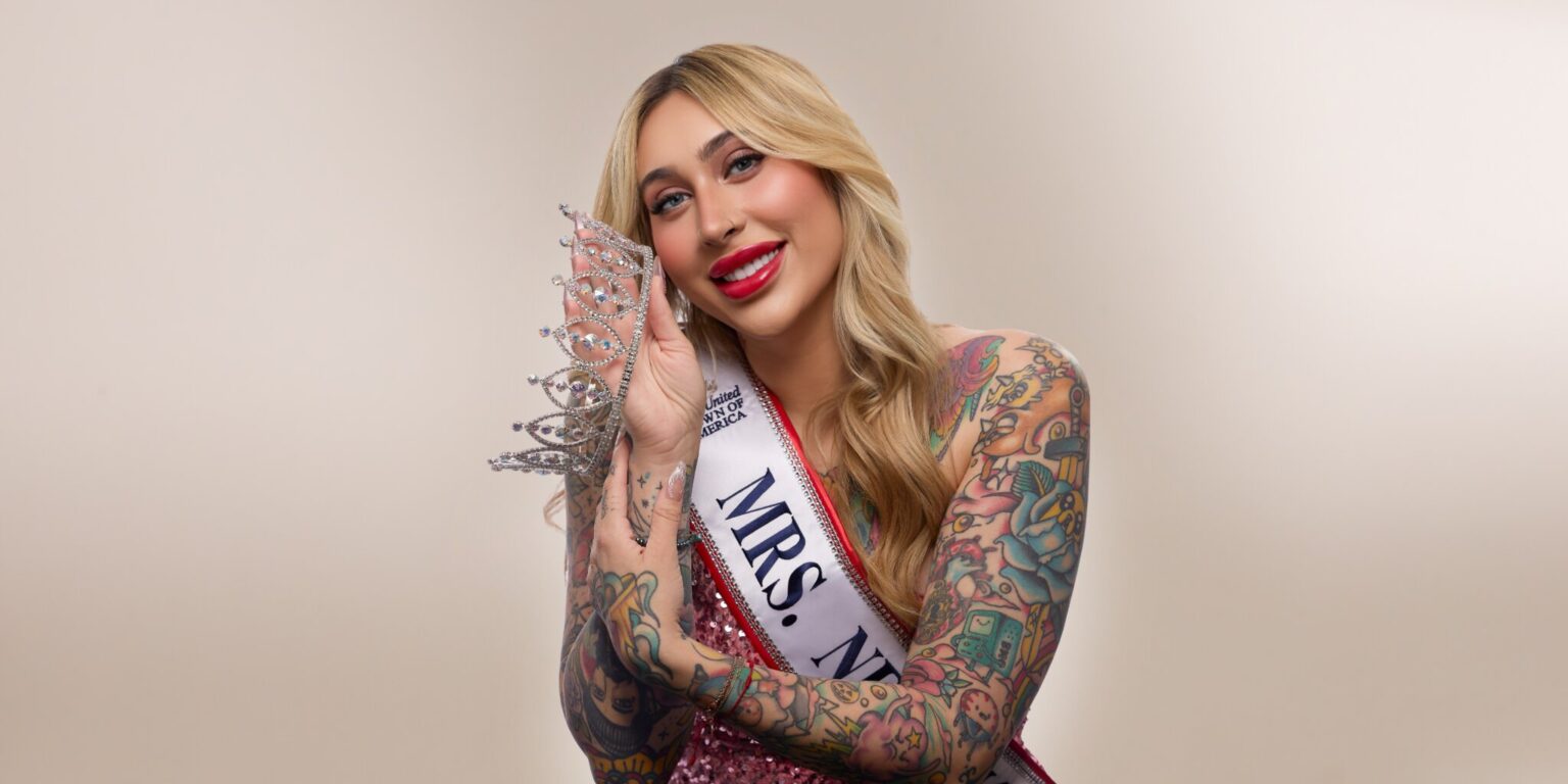 A smiling woman with long blonde hair and vibrant tattoos wears a sparkling crown and "Mrs. NY" sash, exuding confidence and joy against a neutral background.