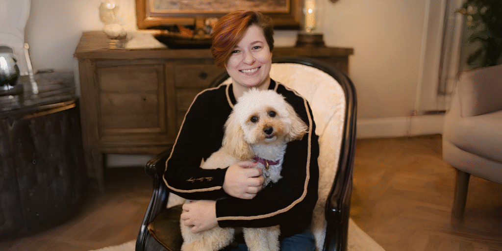 Gabrielle Wanek Redefines Modern Fantasy with Songs of Ash and Dawn A woman sitting with a dog in a cozy chair, smiling.