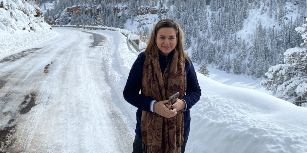 Woman holding phone on snowy mountain road with forested hills and winter landscape behind her.