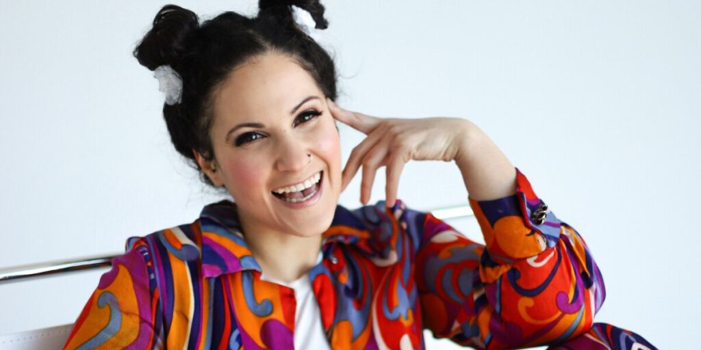 Smiling woman with dark hair in playful buns, wearing a colorful, vibrant shirt with swirling patterns, poses with hand touching her face. Bright and cheerful mood.