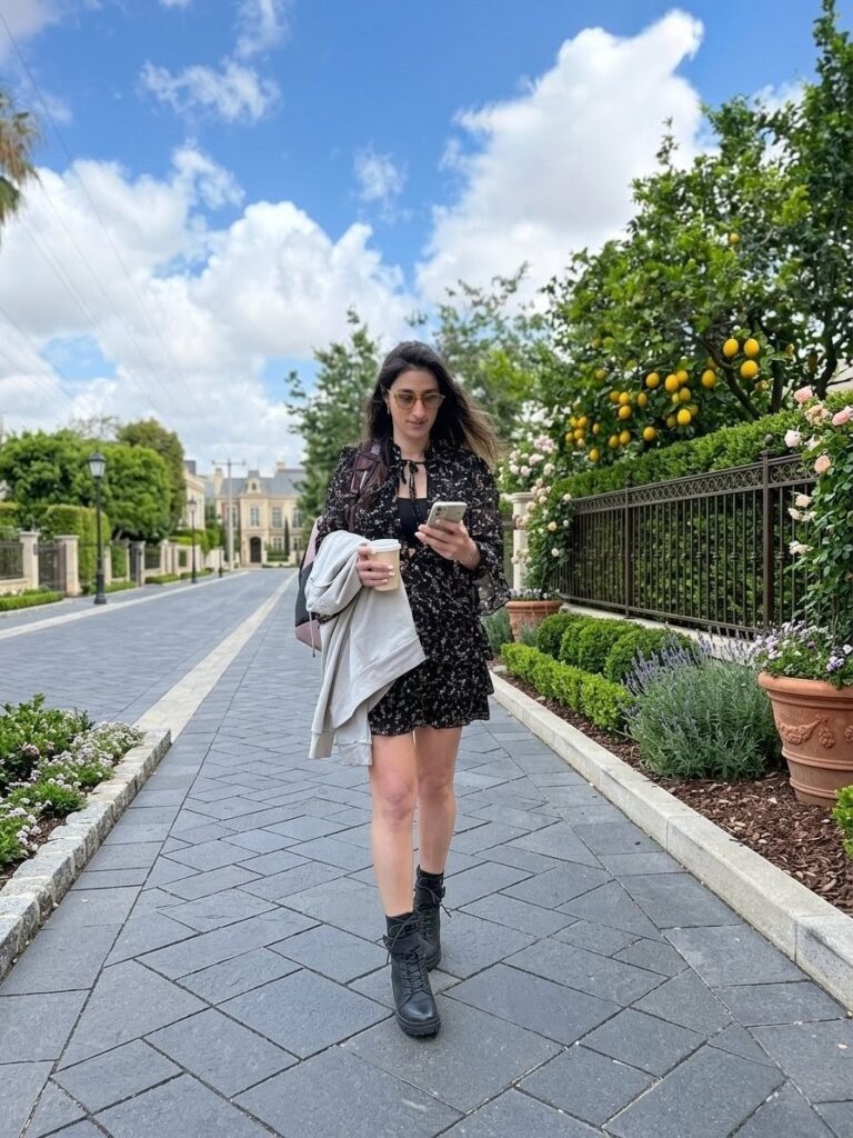 Woman walking down elegant residential street holding coffee and phone beside lemon trees and manicured gardens.