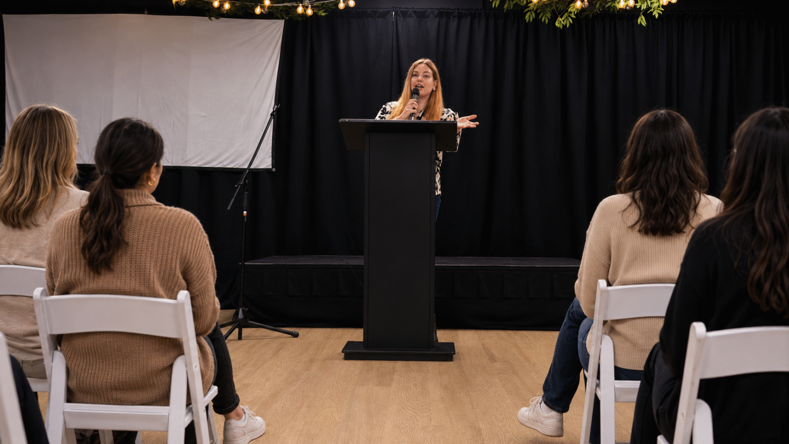 The Girl on the Left: Megan Dirks’ Bold Movement to Redefine Women’s Stories and Empower Their Voice Woman speaking at a podium during a women’s empowerment or storytelling event.
