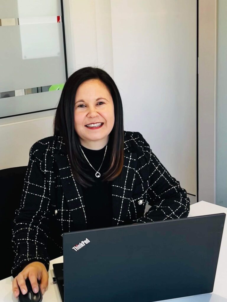 Tracey Franco smiling at her desk, working on a laptop in a professional office.