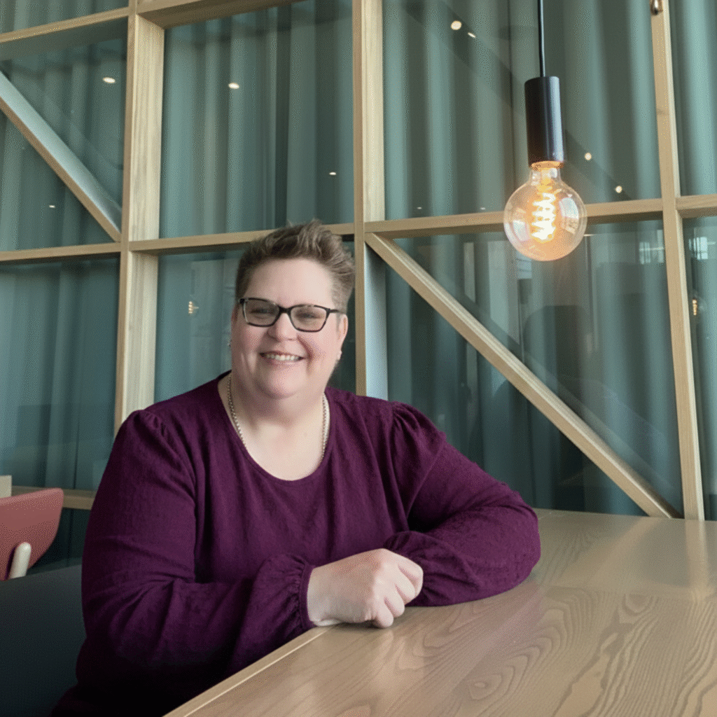 Mickey Forsyth seated at a wooden table, smiling beneath a hanging light in a modern workspace.