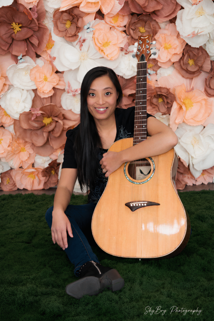 Unbreakable Libby smiles beside an acoustic guitar against a floral backdrop, representing resilience through music.