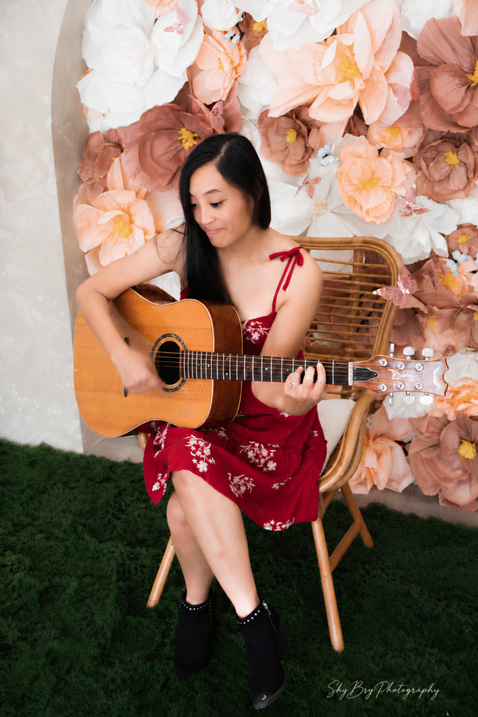 Unbreakable Libby plays acoustic guitar seated before oversized flowers, capturing her passion for songwriting and healing.