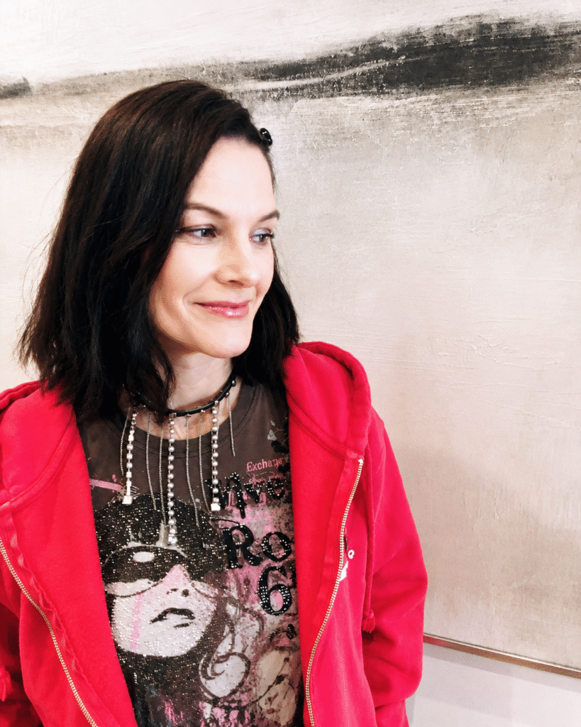 Hapi Reeping smiling indoors, wearing a bright pink hoodie and graphic top against a neutral abstract backdrop.