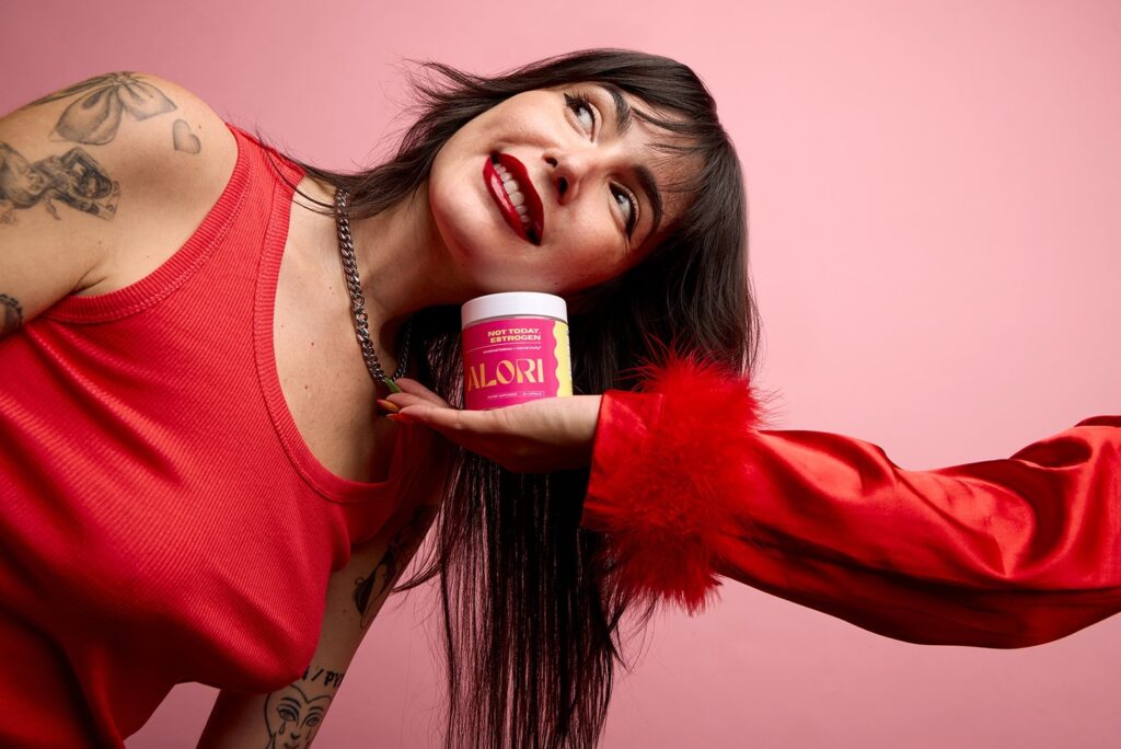 Smiling woman holding Alori supplement container against pink background with red clothing and visible tattoos.