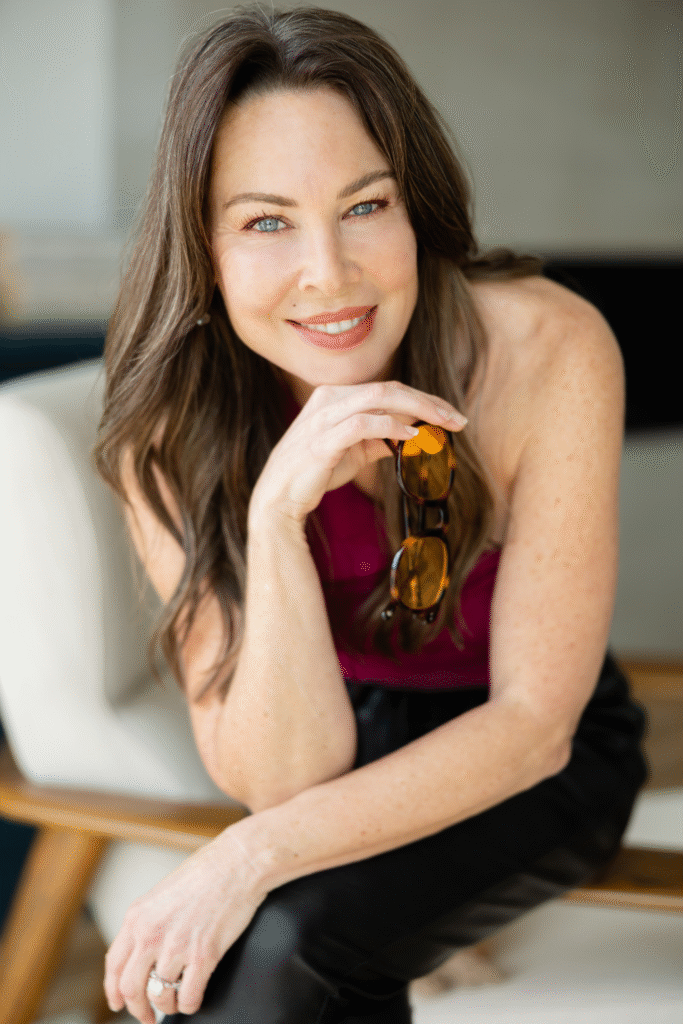 Kim Wheeler smiling while holding sunglasses during an indoor portrait.