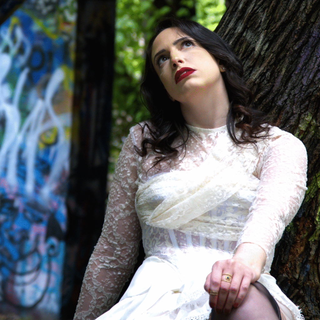 Woman in a white lace dress leaning against a tree in a wooded area.