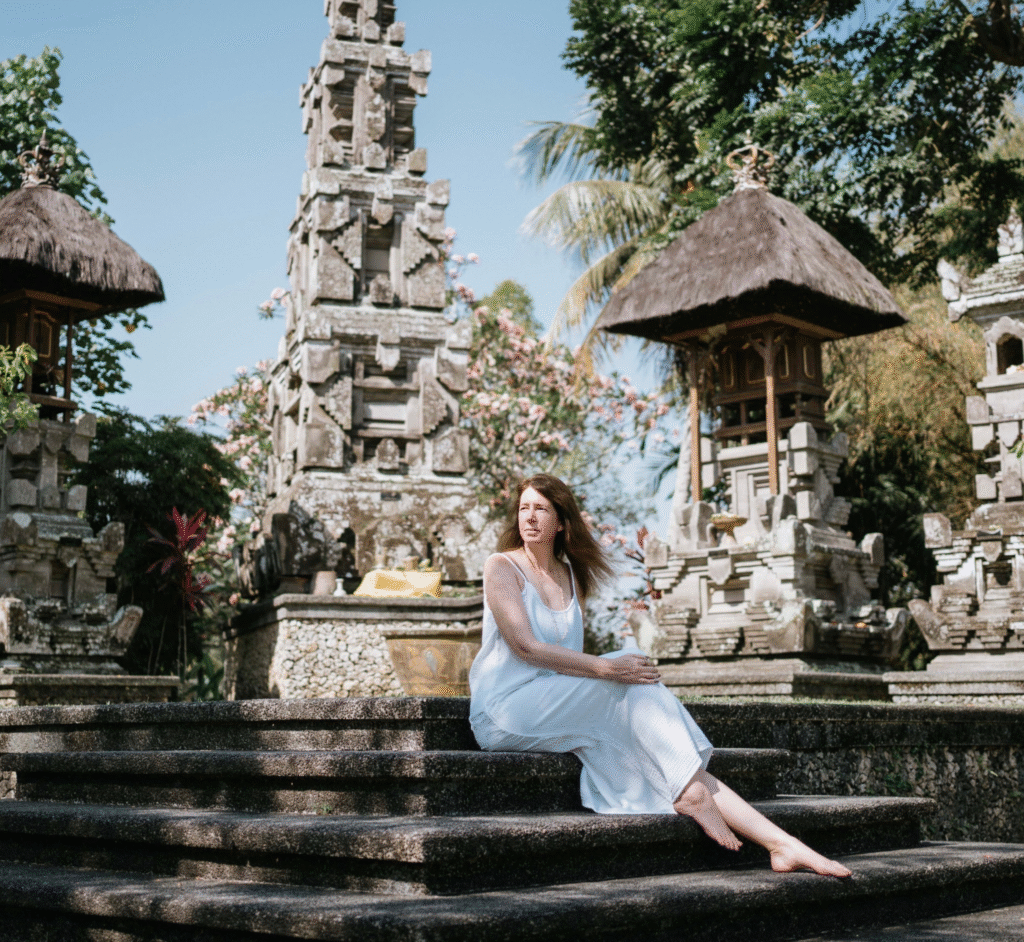 Woman in a white dress seated on temple steps surrounded by traditional Balinese stone architecture.