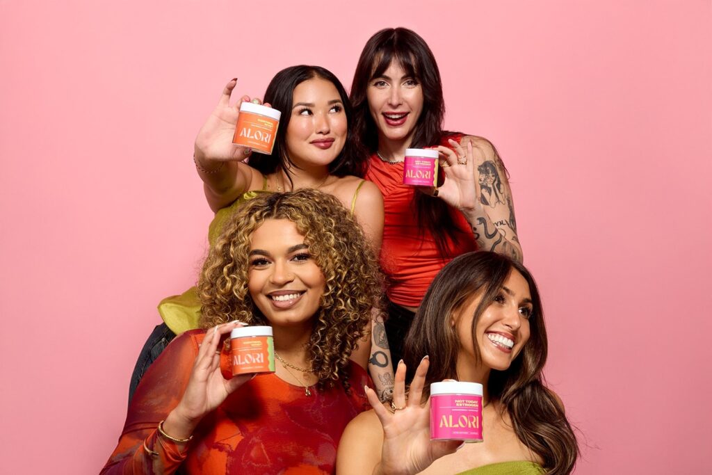 Four women holding Alori supplement containers together, smiling against a pink background.