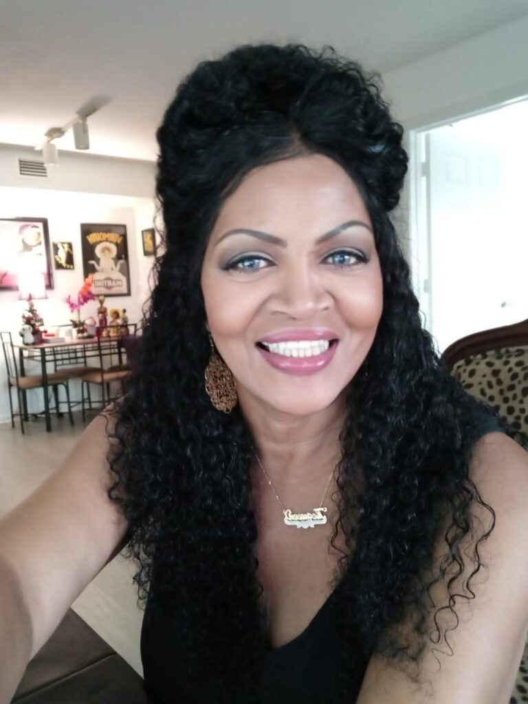 The Psychic Queen smiling indoors with long dark hair, earrings, and a black top.