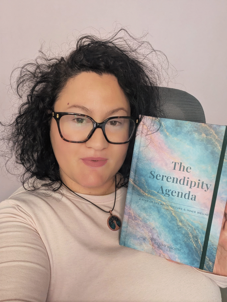 Penny Naoum holding The Serendipity Agenda beside her face while seated indoors.