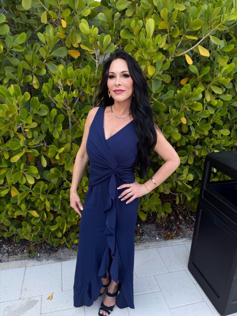 Michele Hunter wearing a navy jumpsuit outdoors in front of green foliage.